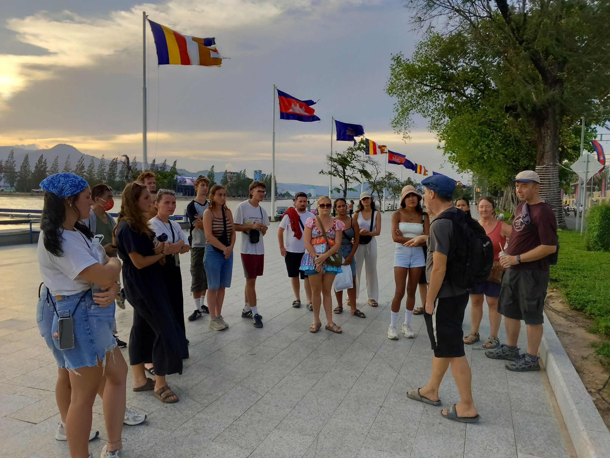 A tour group listening to their guide on the Kampot riverside promenade at sunset, flags flying overhead